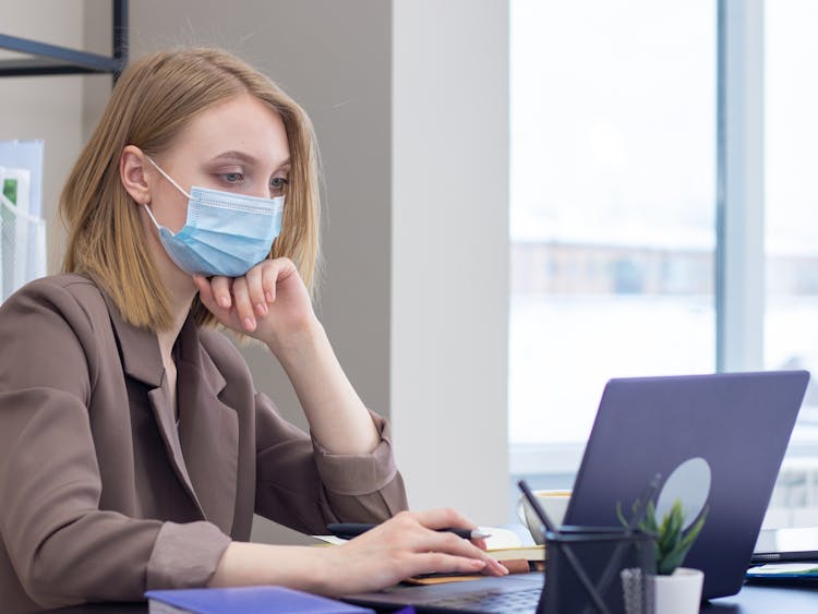 Woman In Brown Blazer Wearing A Face Mask Using A Laptop