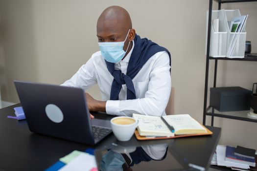 An African American entrepreneur works safely with a laptop and mask in a modern office setting.