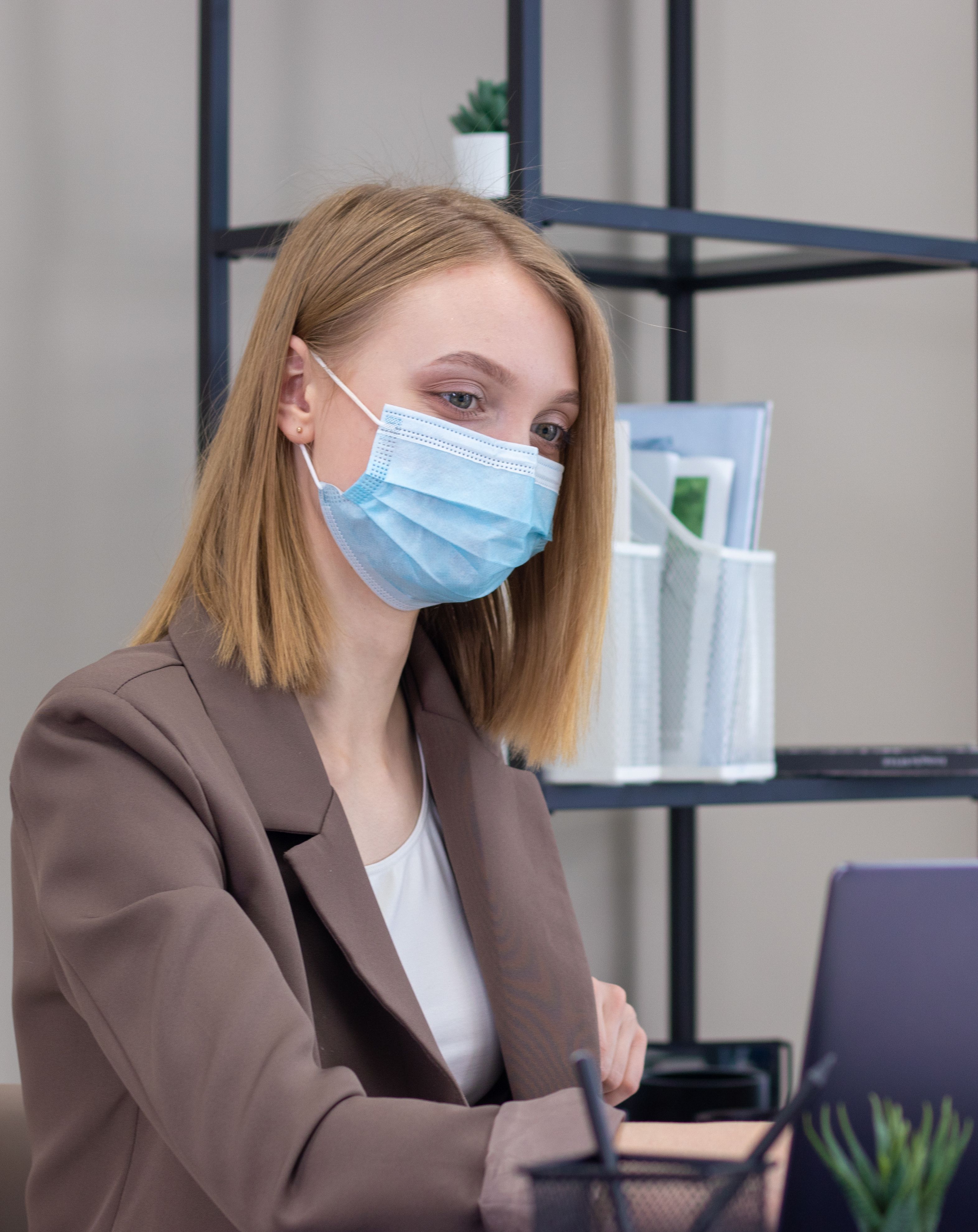 Professional woman in blazer working on laptop, wearing face mask indoors. Modern business setting.