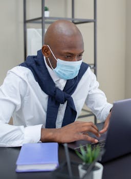 Business professional working on a laptop in office while wearing a protective face mask for health safety.