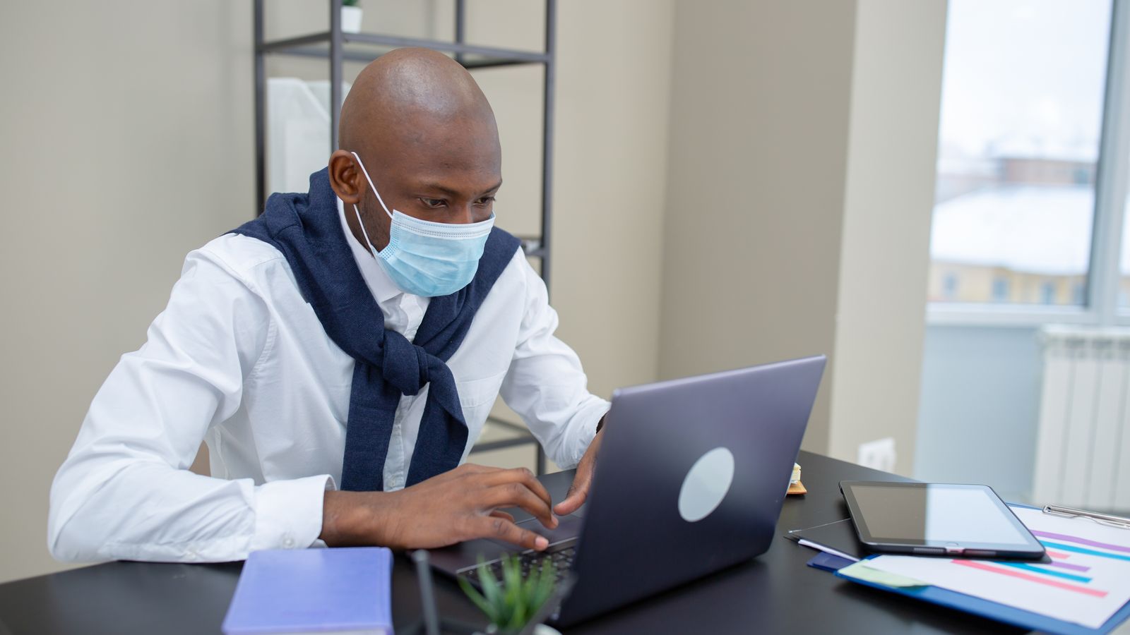 Man wearing a face mask working on a laptop in a modern office setting.