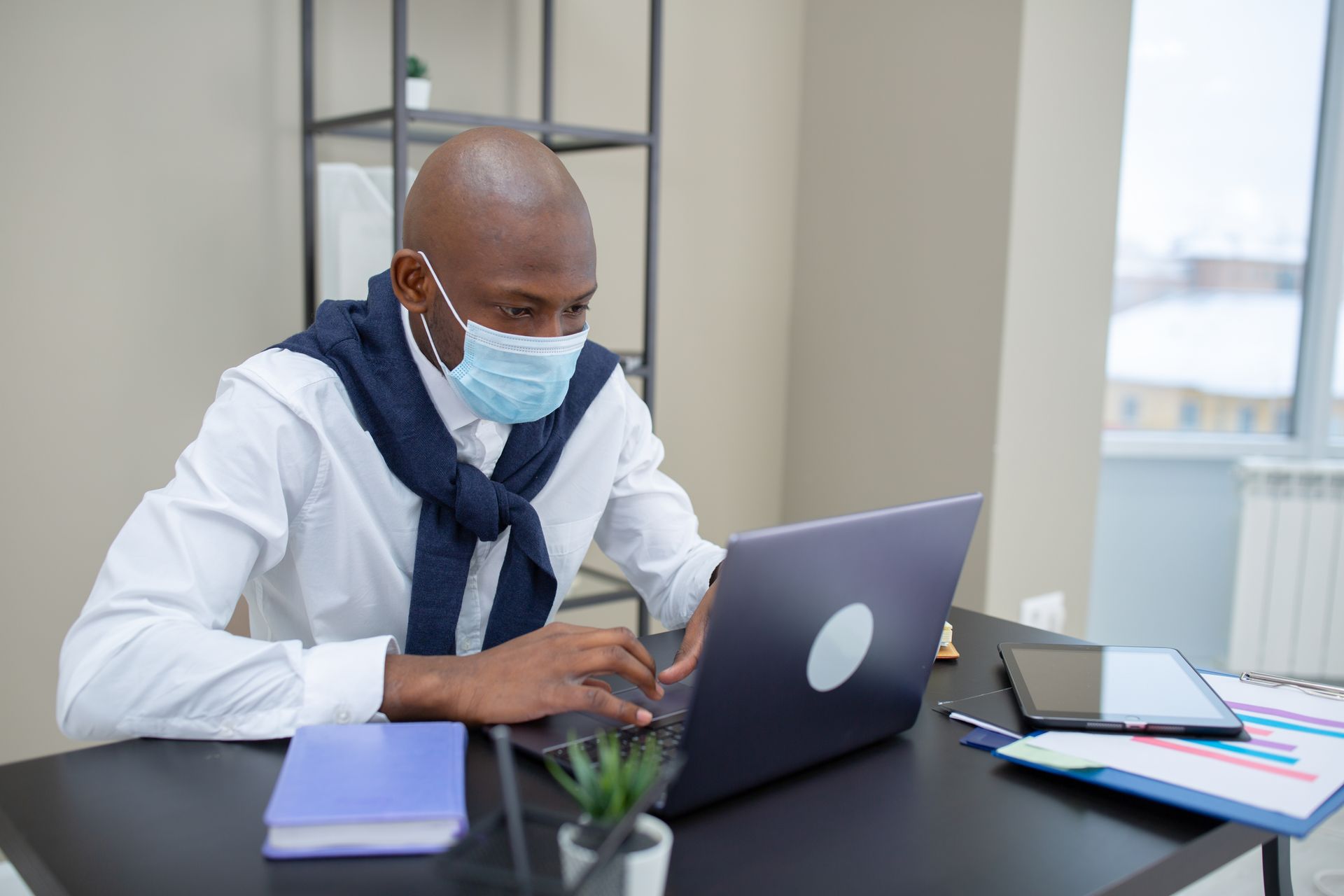 Man wearing a face mask working on a laptop in a modern office setting.