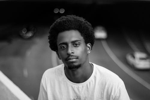 Black and white of young African American male looking at camera on blurred background of asphalt road with automobiles