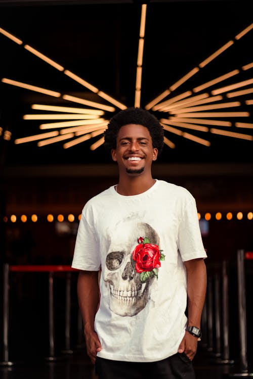 Young glad African American male in stylish T shirt smiling and looking at camera on blurred background of bright lights