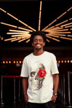 Young glad African American male in stylish T shirt smiling and looking at camera on blurred background of bright lights
