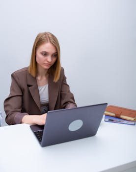 Young businesswoman in a blazer working intently on a laptop at a minimalist office desk.