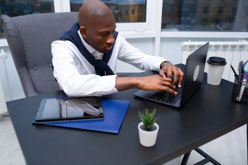 Free Black man in a white shirt using a laptop at a modern office desk. Stock Photo