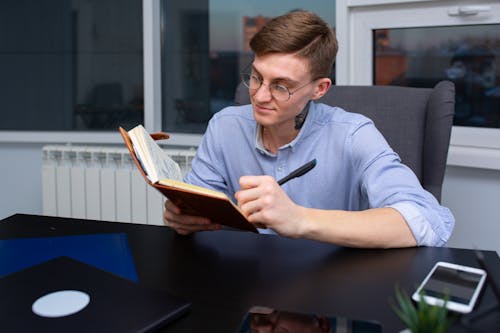 Man in Blue Long Sleeve Shirt Sitting by the Black Table while Writing on Brown Notebook