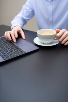 A person's hand holding a coffee cup next to a laptop on a desk, ideal for work-related imagery.