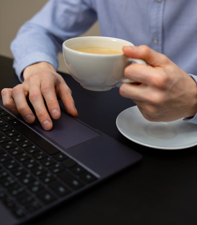 Crop anonymous male manager using touchpad while working on laptop and drinking aromatic cappuccino in morning office