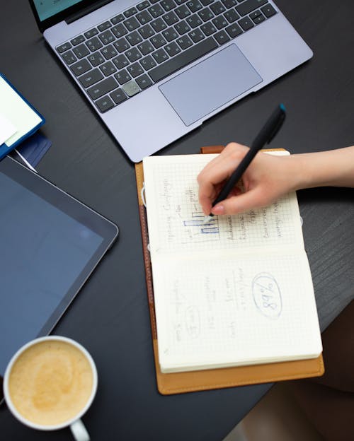 Free A Person Writing on the Graphing Notebook Beside the Laptop and Ipad Stock Photo