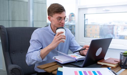 A Man Holding a Coffee Cup While Sitting at the Table with Laptop and Notebook