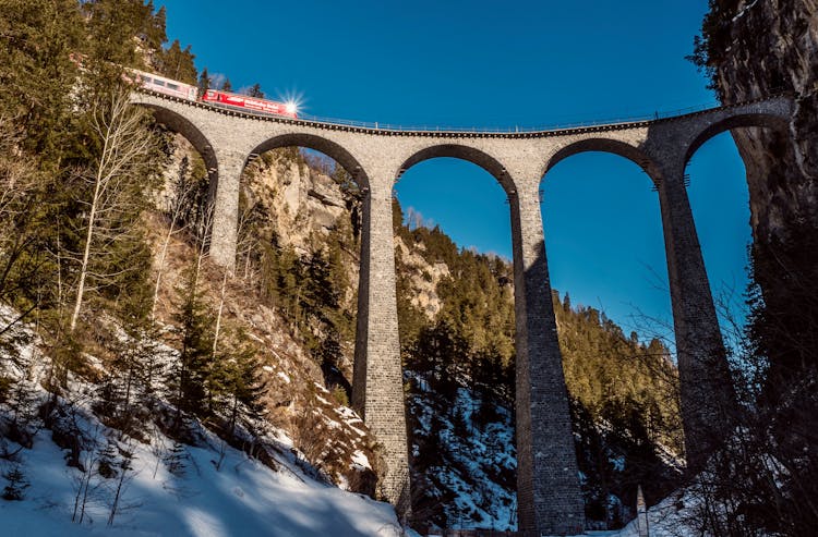 Train Driving On Viaduct Between High Mounts In Winter