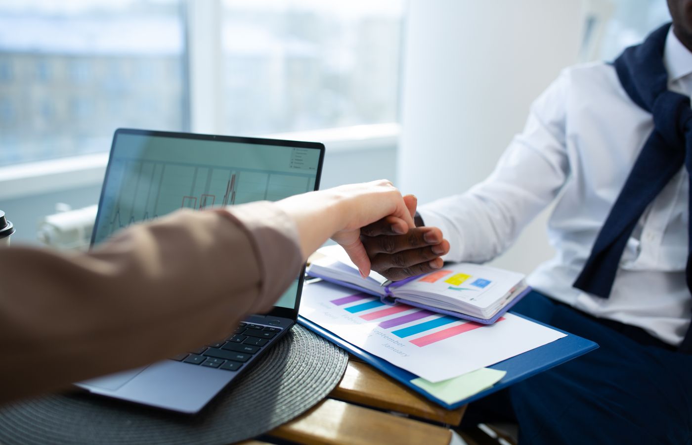 Two business professionals shaking hands over graphs and reports in an office setting.
