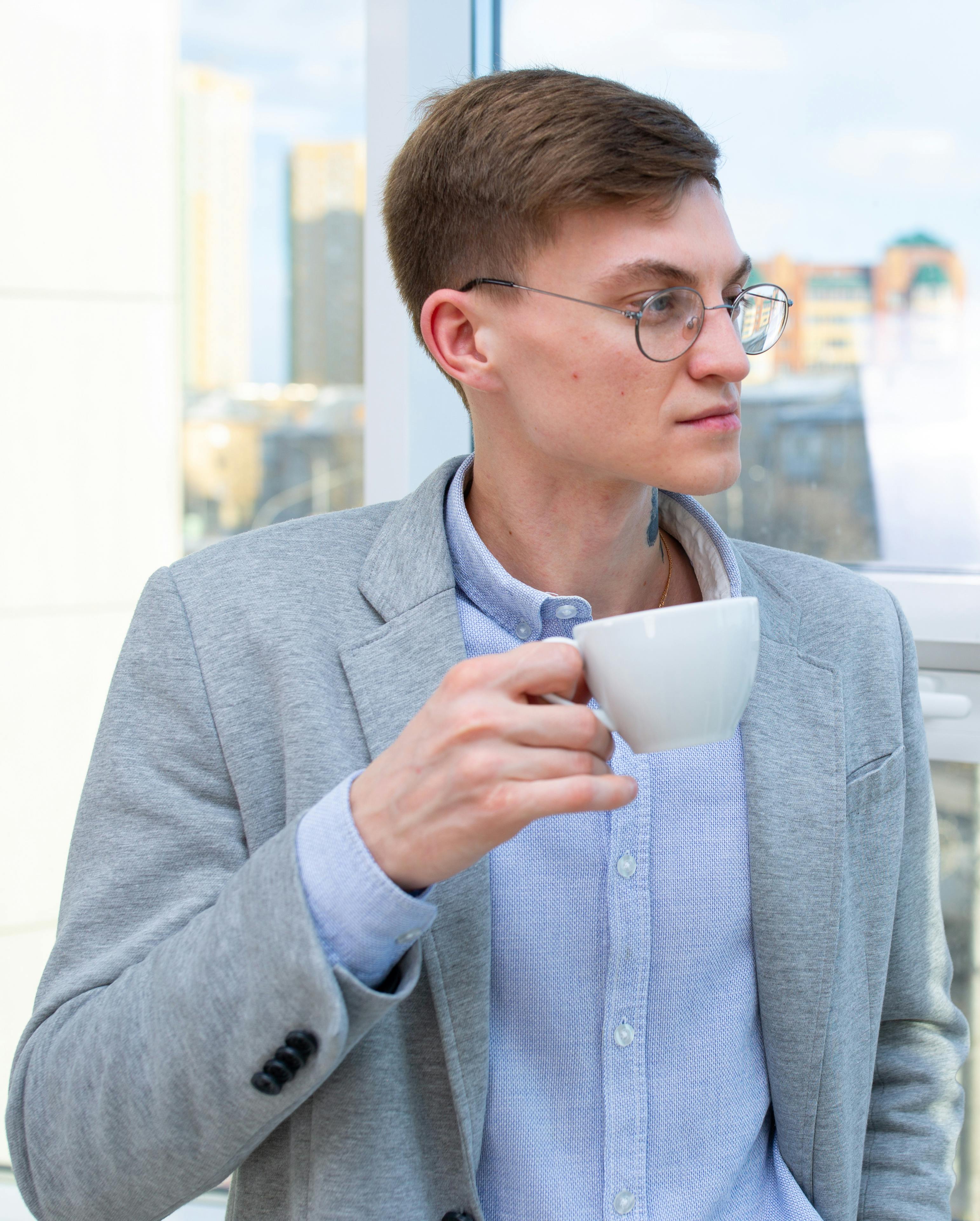 Young professional in office setting holding a coffee mug, wearing glasses and business attire.
