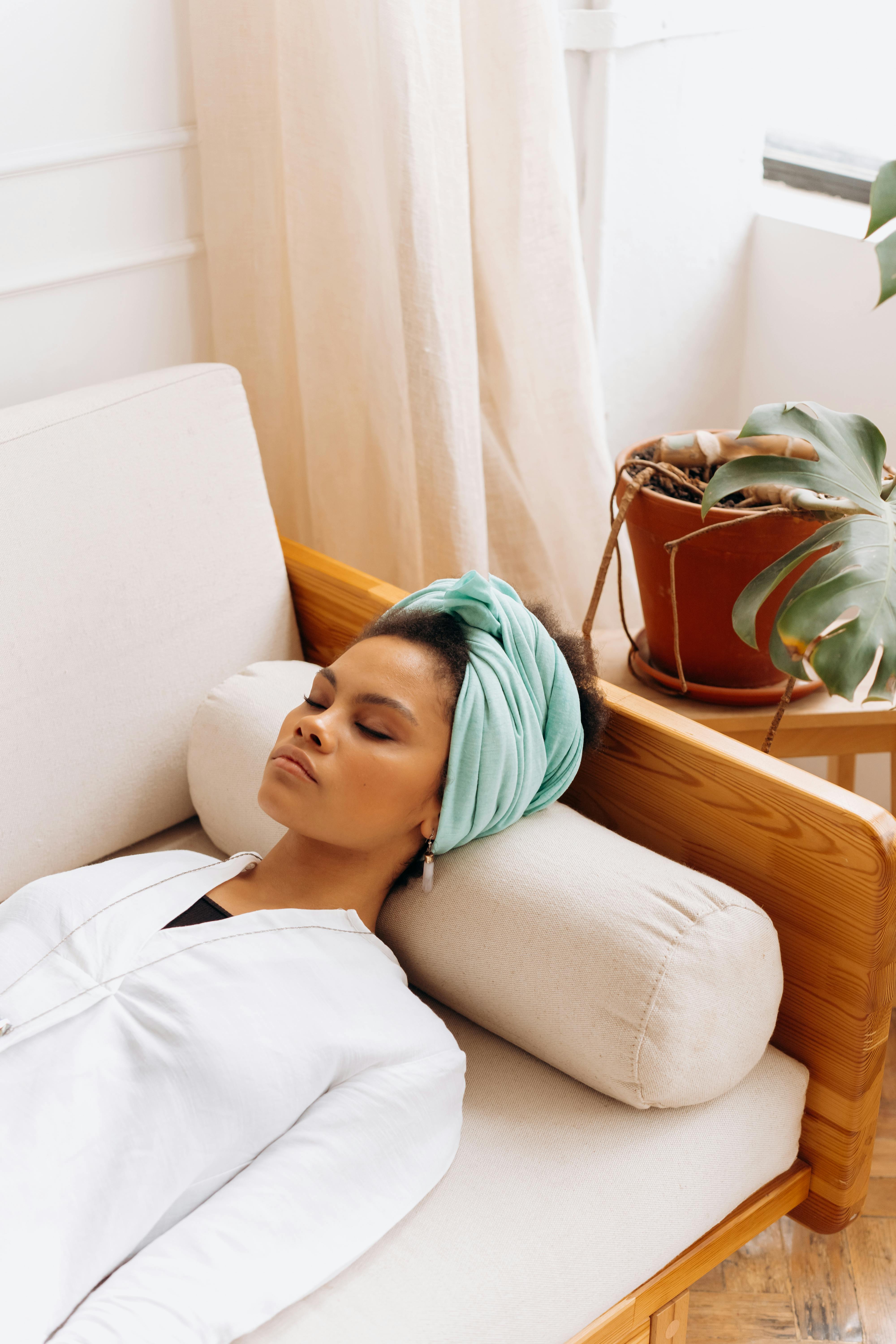 A peaceful adult woman rests on a sofa with eyes closed, wearing a headscarf and white blouse.
