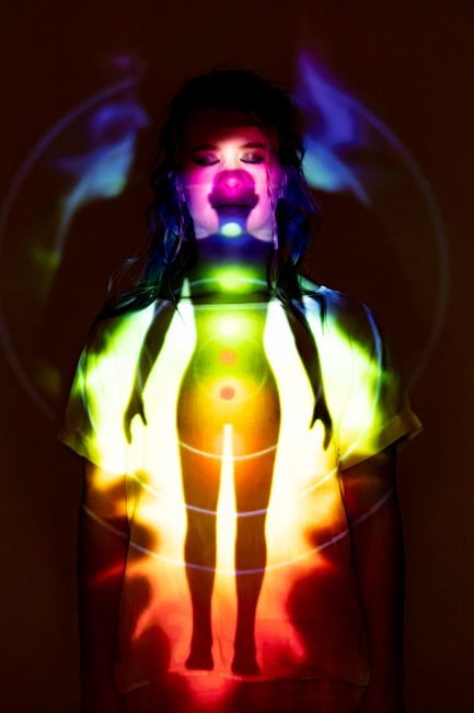 A woman stands in a dark room with a colorful chakra projection on her body.