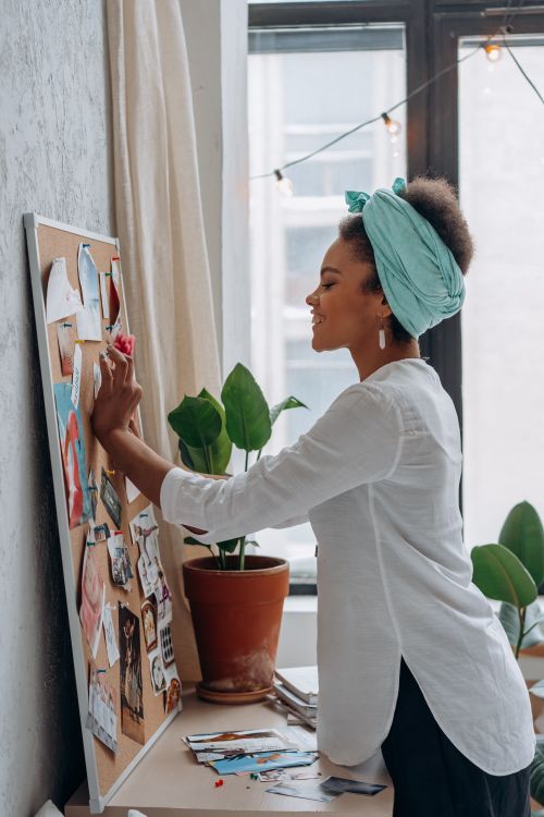 Free Side view of a smiling woman organizing a cork board with photos in a plant-decorated room. Stock Photo