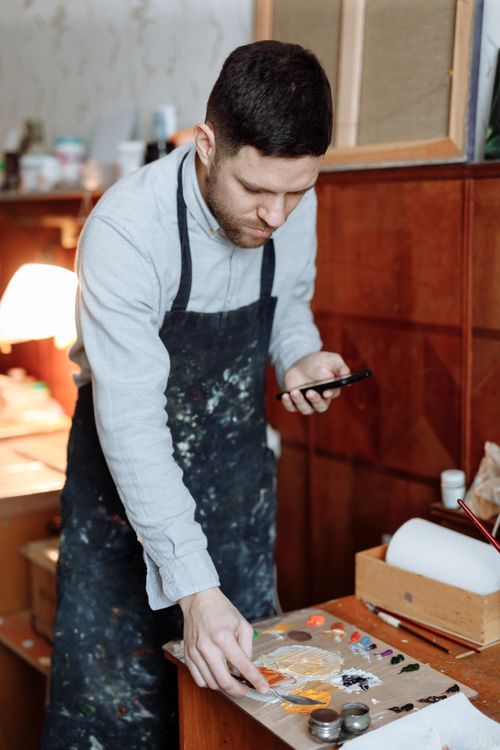 Free Artist mixing colors on a palette while using a smartphone in a studio. Stock Photo