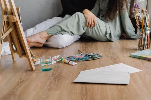 Artists sitting on floor with art supplies, paintbrushes, and an easel creating art in a cozy indoor space.