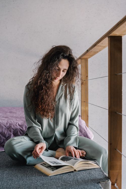 Free Woman sitting on floor in cozy pajamas, reading a book indoors by the railing. Relaxed morning scene. Stock Photo
