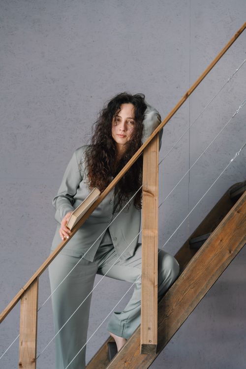 Free Woman in a relaxed pose leaning on wooden staircase indoors, wearing a grey suit. Stock Photo