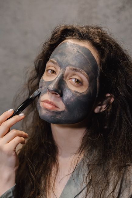 A woman applies a charcoal face mask to her face in an indoor bathroom setting, promoting skincare and relaxation.