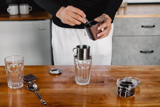 Close-up of hands preparing coffee using a metal dripper on a wooden counter with glass cups.