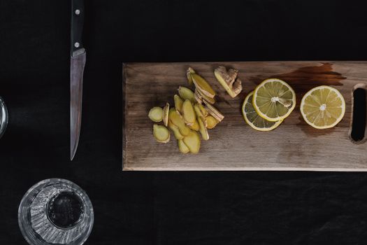 Top-down view of sliced ginger and lemons on a wooden chopping board with knife and glass.