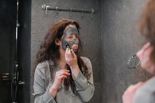 A woman applying a black facial mask while looking in the mirror in a contemporary bathroom.