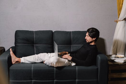 A man in casual attire working remotely on a laptop while relaxing on a comfortable sofa.