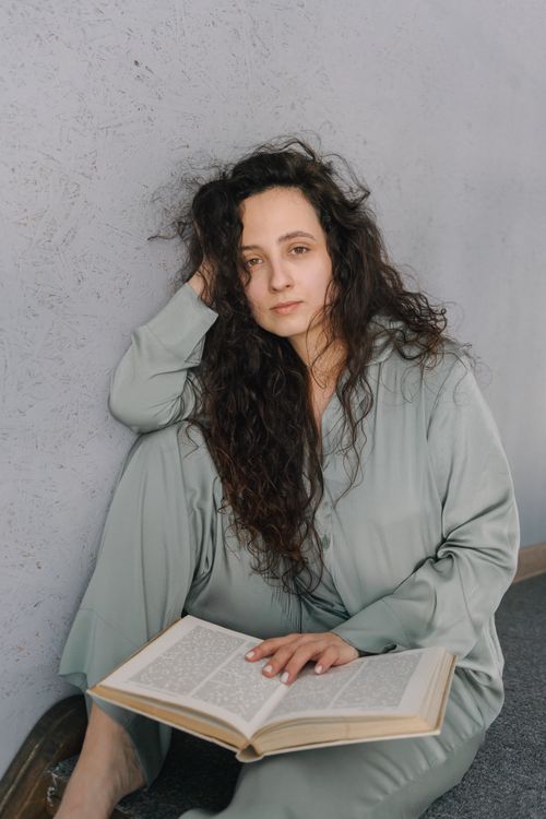 Free Woman in pajamas sitting against a wall, holding a book, with a relaxed and thoughtful expression. Stock Photo