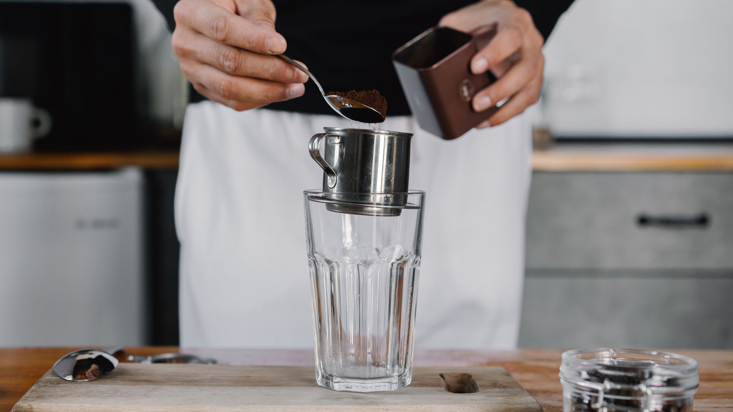 Person preparing coffee using a metal filter over a glass cup on a wooden board.