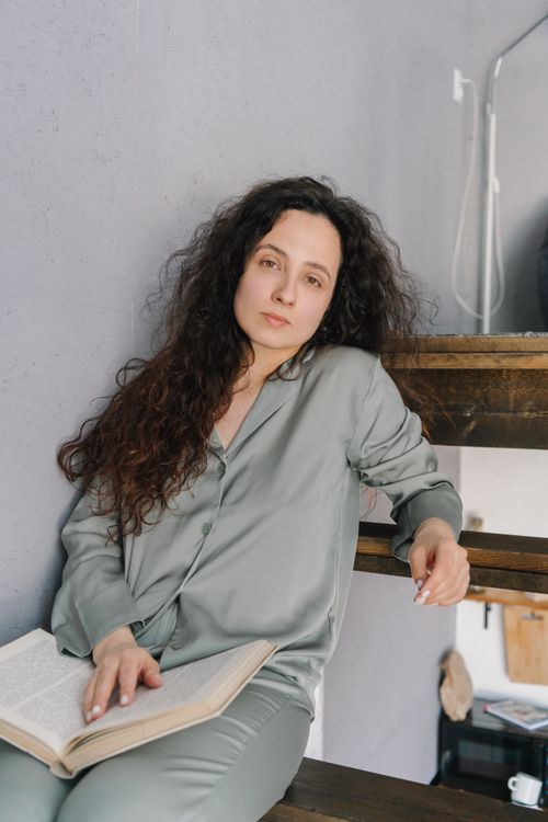 Free Woman with curly hair relaxes on stairs reading a book in cozy pajamas. Stock Photo
