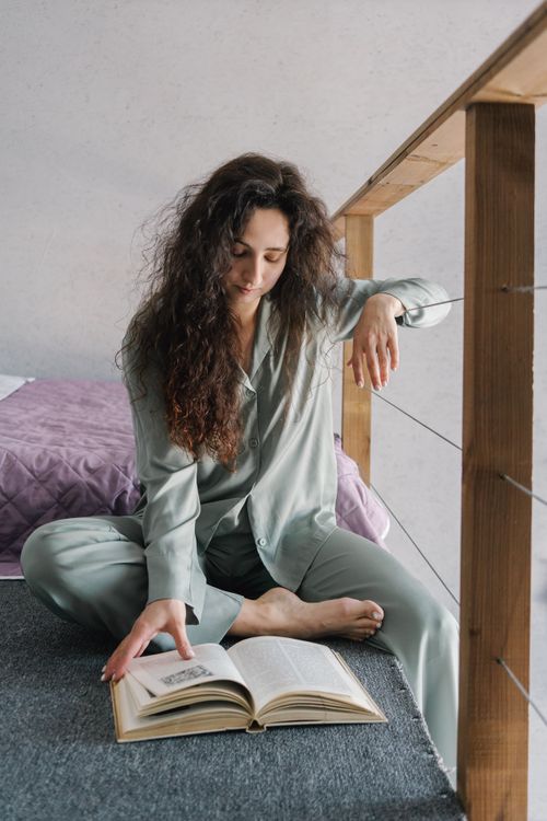 Free Woman in pajamas sitting indoors, reading a book on the floor, enjoying a quiet moment at home. Stock Photo