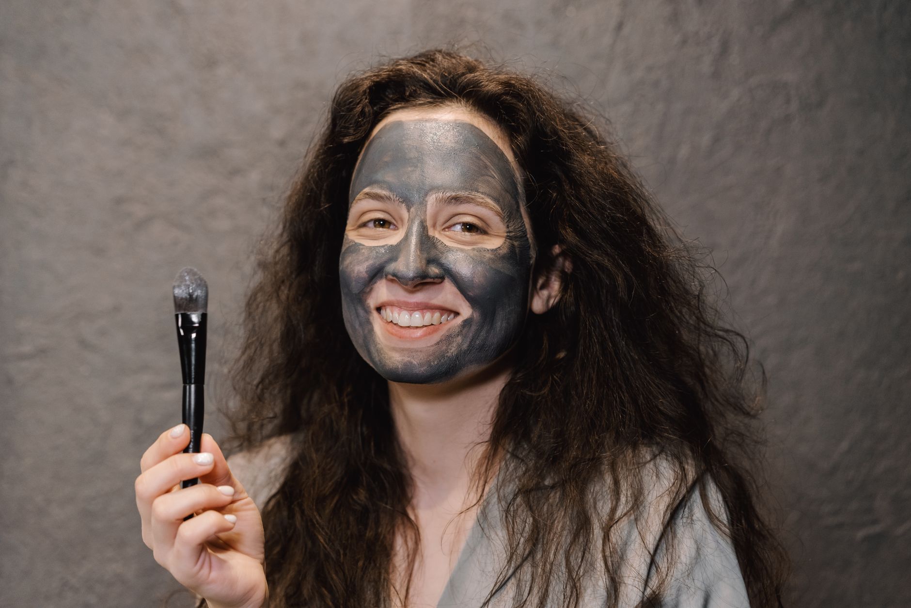 Smiling woman with messy hair applies a gray mud mask using a brush indoors.