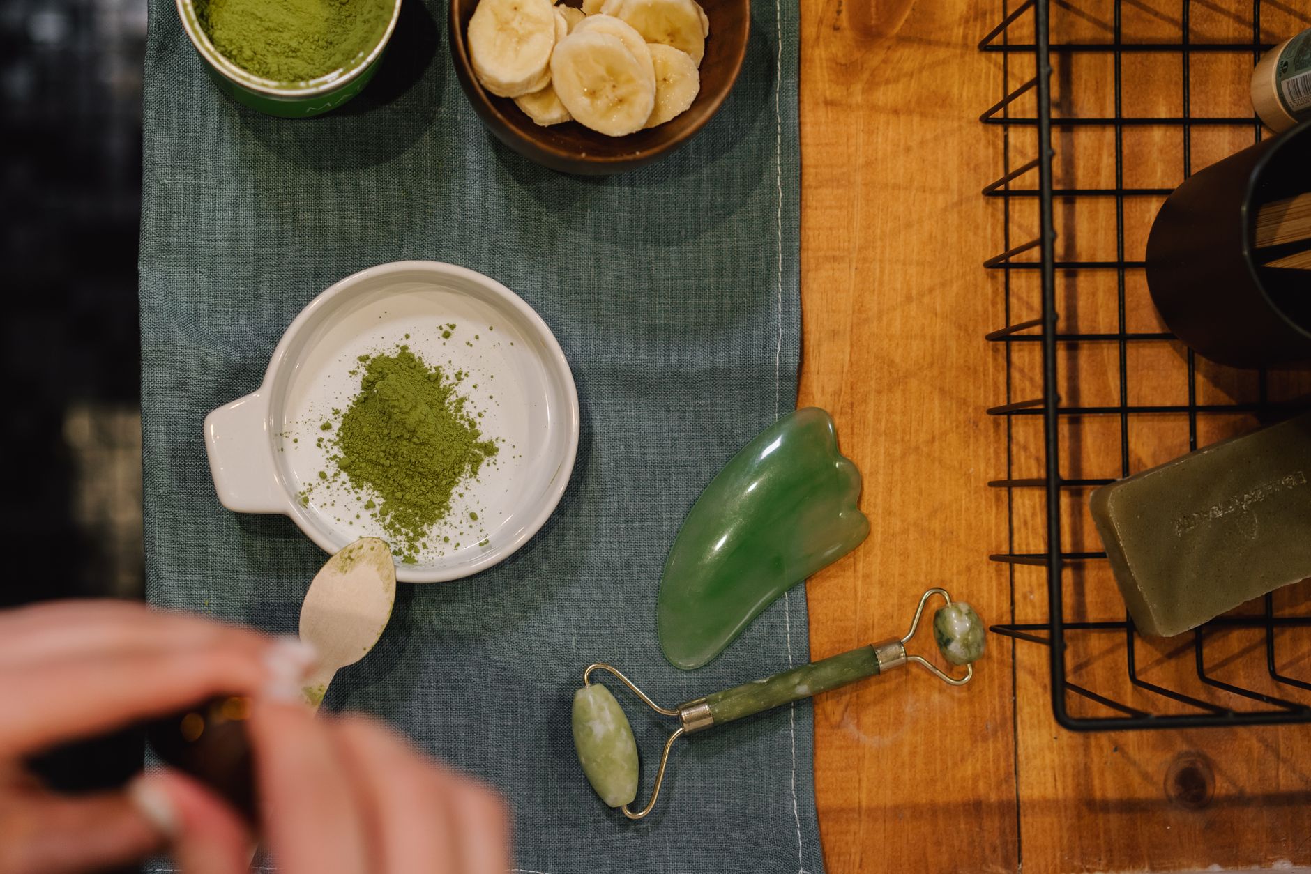 Flat lay of wellness products including jade roller, matcha, and banana slices on wooden table.