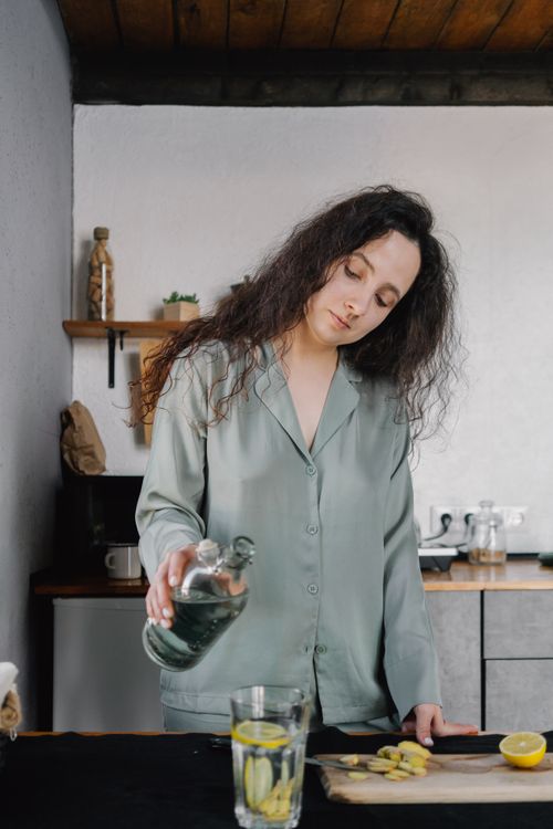 Free A woman pours water into a glass with lemon and ginger, highlighting a home wellness routine. Stock Photo