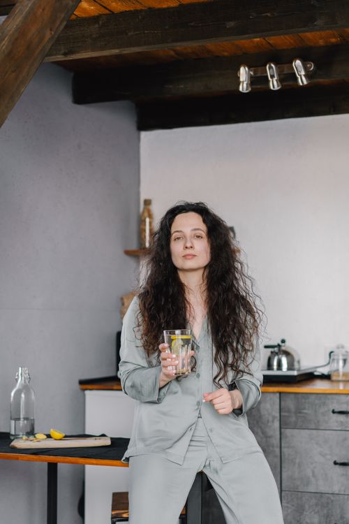 Free Adult woman in pajamas sitting indoors holding a lemon water glass, serious expression. Stock Photo