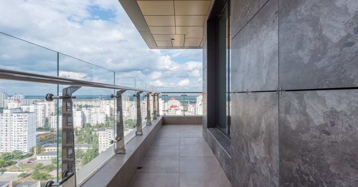 Spacious terrace with glass barrier and metal railings of contemporary apartment in multistory building against cloudy blue sky in city