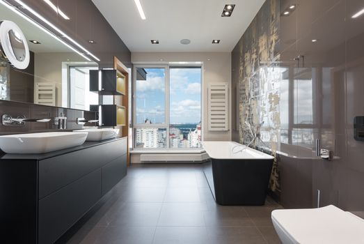 Wooden furniture with bathtub and ceramic sinks and toilet bowl in contemporary master bathroom with big windows in daylight