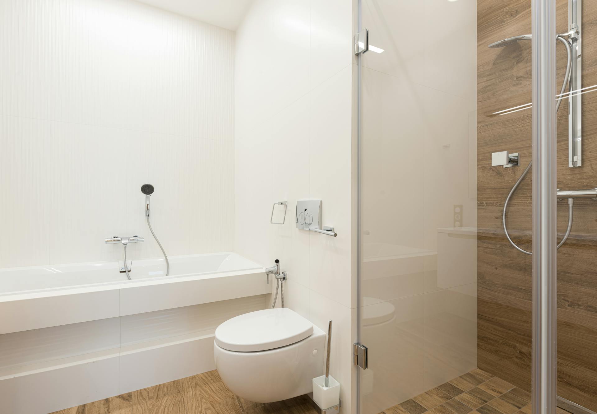 Shower cabin with glass door and ceramic bathtub and toilet bowl in spacious light bathroom with wooden floor