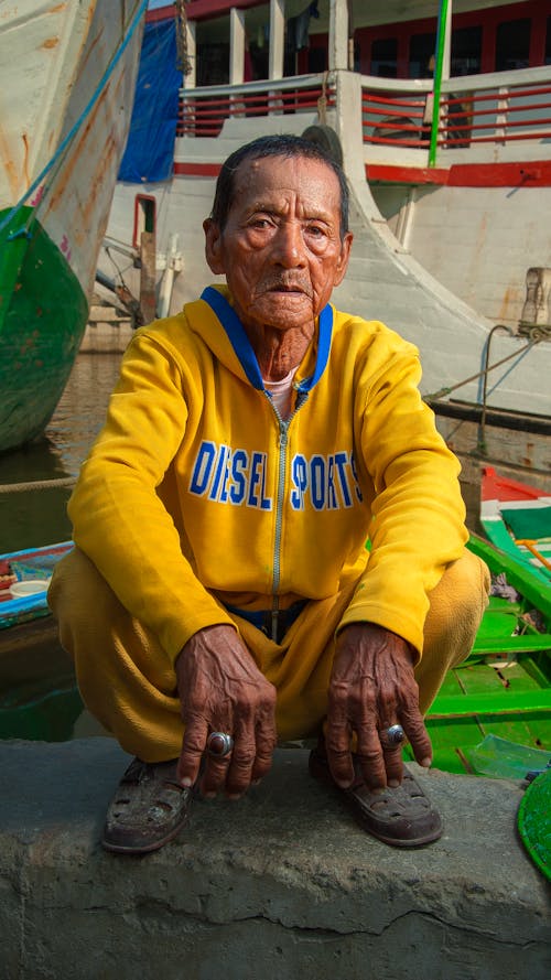 Elderly ethnic male in bright apparel with Diesel inscription squatting in city harbor while looking at camera