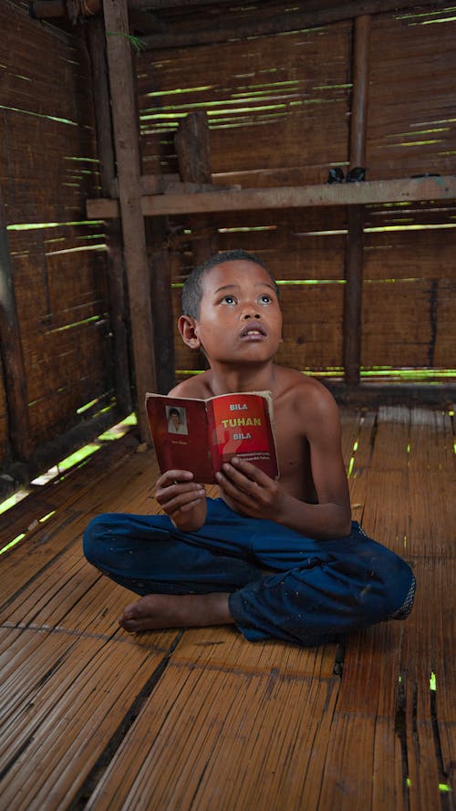 Contemplative ethnic child with textbook looking up while sitting with crossed legs on bamboo floor