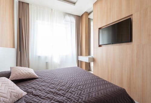 Interior of bedroom in minimalist style with bed and TV set hanging in niche in wooden wall