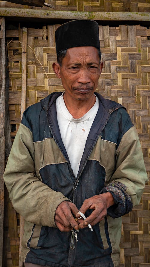 Elderly Asian male with wrinkled face skin and cigarette looking at camera against straw wall