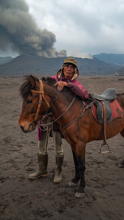 Ethnic male in rubber boots leaning on stallion in bridle while looking away against mountains under cloudy sky