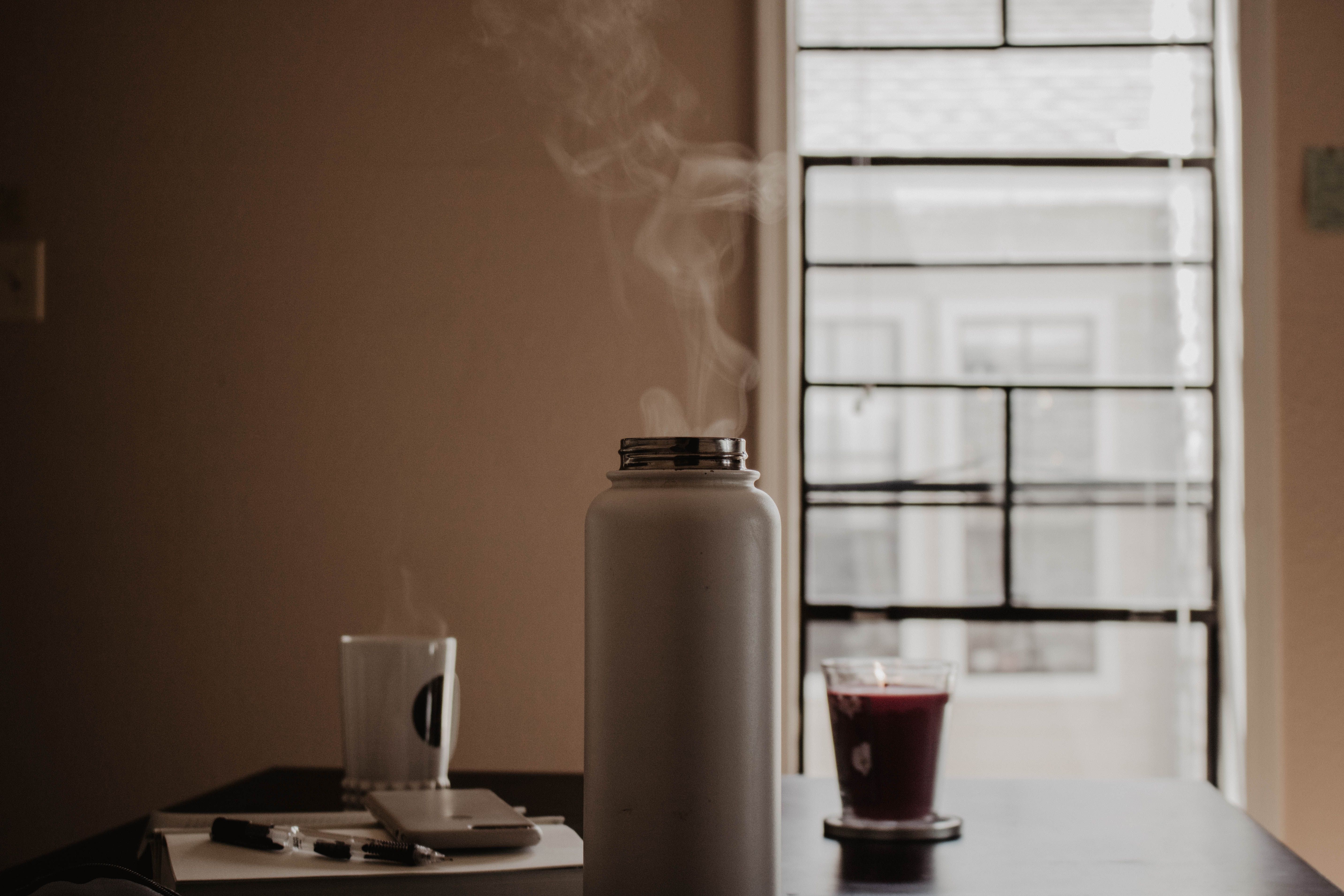 A subtle warm scene showing a steaming thermos, mug, and notebook on a wooden table in an indoor setting.