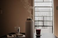 A subtle warm scene showing a steaming thermos, mug, and notebook on a wooden table in an indoor setting.