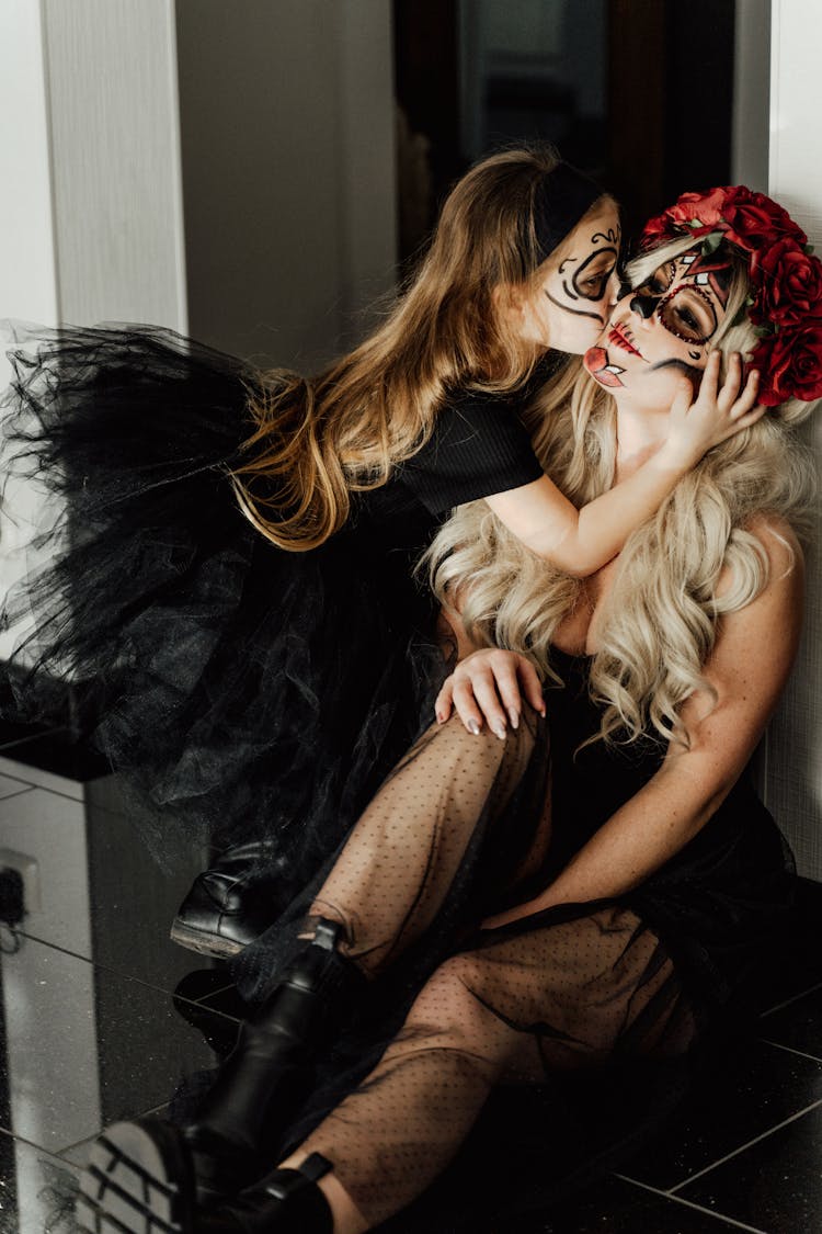 A Girl In Halloween Costumes Kissing The Cheek Of A Woman Sitting On The Floor